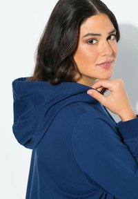 Žena s tmavými vlasy se otáčí k fotoaparátu, drží kapuci svého modrého mikinového svetru poblíž brady na jednobarevném pozadí.