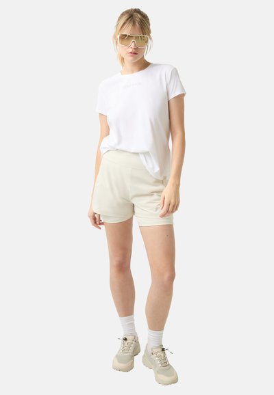 Bílá tričko s jemným logem, béžové šortky, světlé ponožky a masivní béžové tenisky, doplněné o oversized sluneční brýle.