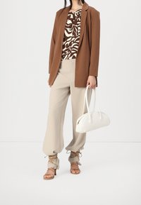 Brown tailored blazer over a brown and cream leaf-pattern top, beige wide-leg pants, and white curved handbag with tan strappy heels.