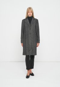 Cappotto grigio a zig zag con rever a scendere, due bottoni e tasche laterali; indossato sopra un dolcevita nero e abbinato a pantaloni scuri.