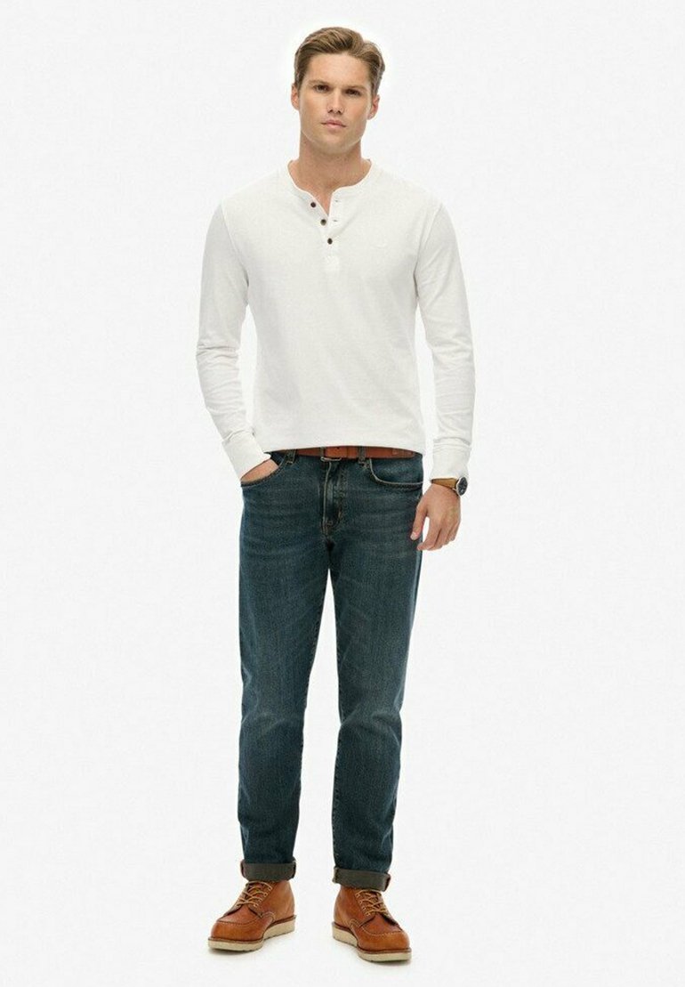 Weißes Langarm-Henley-Shirt mit drei Knöpfen, kombiniert mit dunkelblauen Jeans und braunen Schnürschuhen. Klassisches und vielseitiges Design.
