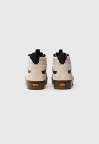 Beige høyhælte Vans-sneakers med svarte detaljer og brune såler, vist fra baksiden på en ensfarget hvit bakgrunn.