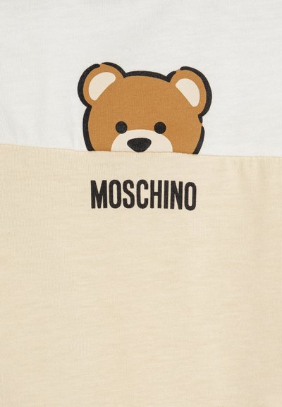 Camiseta de manga corta con un gráfico de un oso pardo en la sección superior blanca y una sección inferior beige claro, con "MOSCHINO" impreso en negro.