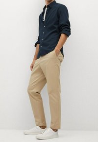 Uomo che indossa una camicia azzurra a bottoni, pantaloni beige e sneakers bianche, in piedi con una mano in tasca contro uno sfondo neutro.