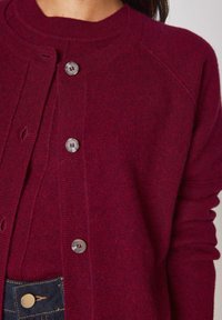 Primo piano di una persona che indossa un cardigan bordeaux con bottoni e un maglione coordinato, abbinati a jeans di denim blu scuro.