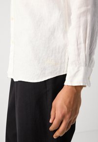 Chemise blanche en lin à manches longues avec un devant boutonné et des poignets, mettant en avant un détail d'étiquette brodé près de l'ourlet. Associée à un pantalon sombre.