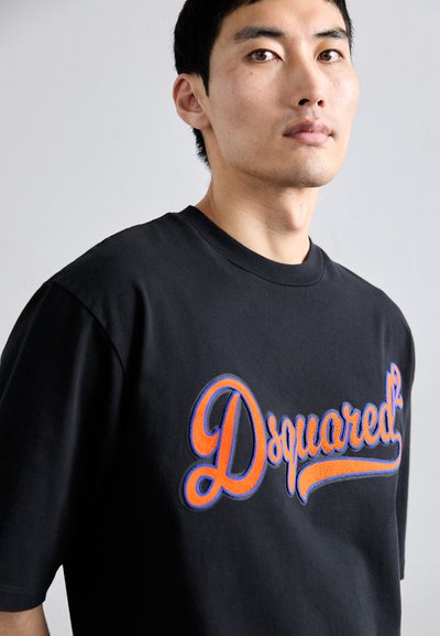 Jonge man in een zwart T-shirt met een vet oranje en blauwe "Dsquared²" logo, staand tegen een effen lichtgrijze achtergrond.