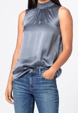 Blouse sans manches en satin gris brillant avec un col montant froncé, portée rentrée dans un jean en denim bleu, une main dans la poche.