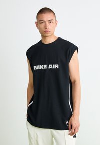 Mlad moški v črni majici brez rokavov Nike Air in belih hlačah, stoji pred enostavno svetlo ozadje in gleda naprej.