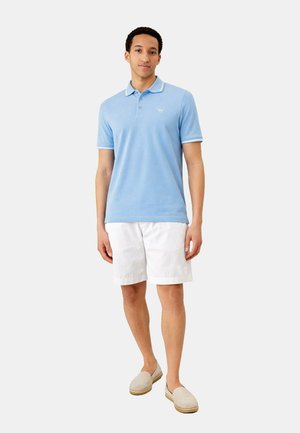 Junger Mann steht, trägt ein hellblaues Poloshirt, weiße Shorts und beige Slipper, vor einem schlichten grauen Hintergrund.