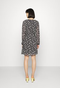 Vestido preto floral com mangas longas, apresentando uma cintura ajustada, um corte ligeiramente em A e padrões de flores brancas. Combinado com sapatos de salto bloco amarelos.