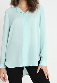 Blusa larga de manga larga en aqua claro, hecha de tela transparente, con cuello de camisa, tapeta de botones y dobladillo curvado, combinada con pantalones negros.