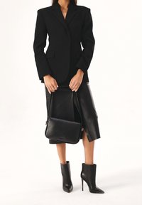 Nainen, joka on pukeutunut mustaan bleiseriin, mustaan nahkahameeseen, jossa on etureikä, mustiin korkokenkiin nilkassa, ja pitää mustaa nahkaista käsilaukkua kahvoista.