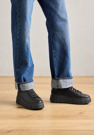 Persoon met opgerolde blauwe jeans en zwarte plateau sneakers staand op een houten vloer tegen een effen muur.