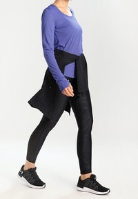 Lila långärmad tröja, svarta leggings med texturerade mönster och svarta träningsskor. En svart jacka knuten runt midjan.