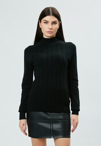 Maglione nero lavorato a maglia con scollo alto e motivo a treccia testurizzato. Abbinato a una mini gonna in pelle nera con cerniera centrale.