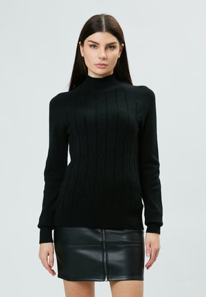 Schwarzer, gestrickter Pullover mit hohem Kragen und strukturiertem Zopfmuster. Kombiniert mit einem schwarzen, ledernen Minirock mit zentralem Reißverschluss.