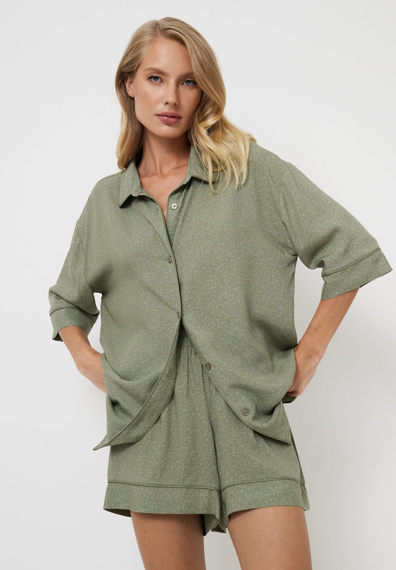 Camisa de botón de manga corta en verde oliva con ajuste relajado y bolsillos delanteros, combinada con pantalones cortos a juego que presentan un patrón sutil.