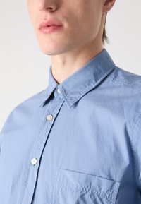 Lichtblauw overhemd met knopen, gemaakt van een soepele stof. Kenmerkt zich door een kraag, een borstzak en parelachtige knopen met gestikte details.