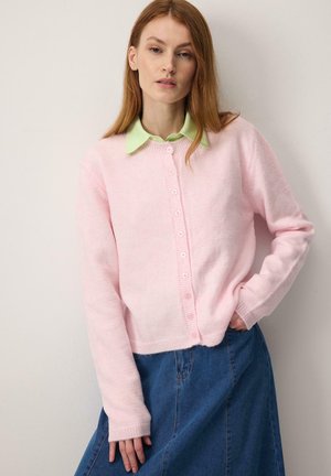 Donna con lunghi capelli rossi, indossa una camicia verde chiaro con colletto, un cardigan rosa pallido abbottonato e una gonna di jeans blu, in piedi contro un muro bianco.
