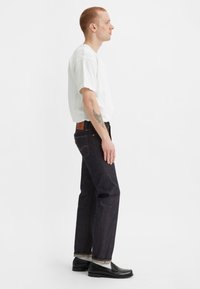 Vaqueros de mezclilla oscuro con corte recto, costuras naranjas y un parche de piel en la parte posterior. Combinados con una camiseta blanca lisa y zapatos negros.