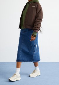 Henkilö kävelee, päällään valkoiset lenkkarit, valkoiset sukat, sininen farkkumidi-hame, jossa on tasku, oliivinvihreä huppari ja ruskea takki.