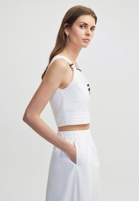 Witte cropped top met zwarte accenten, voorzien van geknoopte bandjes en een textuurafwerking. Model draagt het met hoge witte broek.