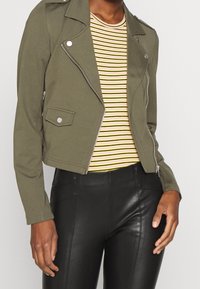 Woman wearing olive green cropped biker jacket, yellow and black striped shirt, and high-waisted black leather pants, standing with hands relaxed.