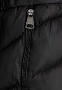 Schwarze Pufferjacke mit einem auffälligen Reißverschluss und einem metallischen Ziehgriff sowie strukturiertem Stoff, der gesteppten Abschnitte für Isolation zeigt.