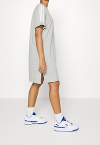 Grå jersey t-shirt klänning med korta ärmar, utrustad med tre vita ränder på axeln och en sidoflits. Kombinerad med vita sneakers.