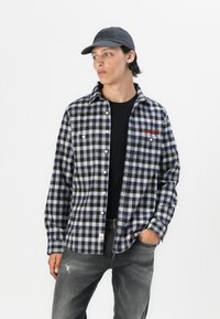 Ruitjes-overhemd in marineblauw en grijs, gemaakt van een textuurstof, met twee borstzakken en een rode logodetail. Gedragen over een zwart shirt en grijze jeans.