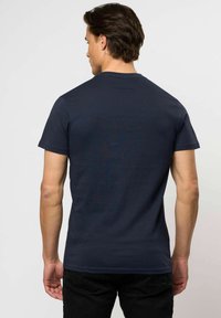 Egy férfi háttal áll, sima sötétkék, rövid ujjú pólót és fekete nadrágot visel, semleges háttér előtt.