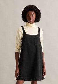 Zwarte denim jurk met een vierkante halslijn, voorzien van een voorzak en verstelbare banden, gelaagd over een lichtbeige coltrui.