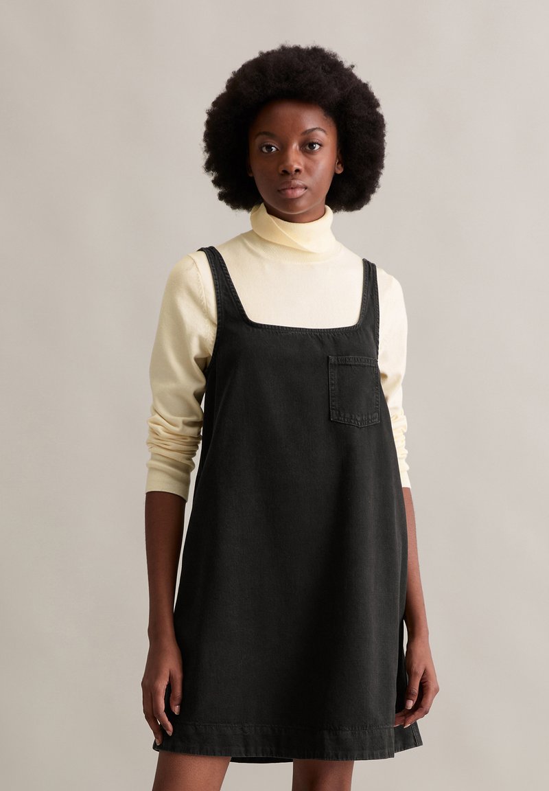 Zwarte denim jurk met een vierkante halslijn, voorzien van een voorzak en verstelbare banden, gelaagd over een lichtbeige coltrui.