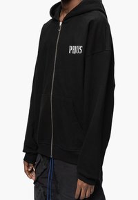 Sweat à capuche noir zippé en tissu doux, avec une poche frontale, des poignets côtelés et le texte « PEQUS » en blanc sur la poitrine.