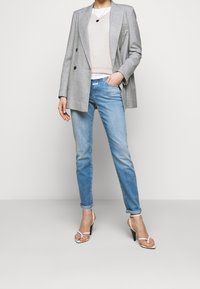 Femme portant un blazer croisé gris clair, un haut beige, un jean bleu clair et des sandales blanches à talons hauts, debout avec une main dans la poche.