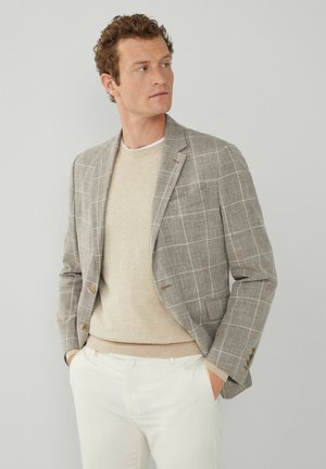 Blazer a cuadros grises con dos botones, llevado sobre un suéter beige claro. Complementado con pantalones de color claro, añadiendo un toque clásico.