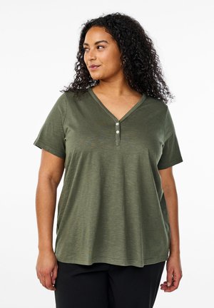 Frau mit lockigem Haar, die ein olivgrünes, kurzärmliges V-Ausschnitt-Shirt mit zwei Knöpfen trägt und vor einem weißen Hintergrund steht.