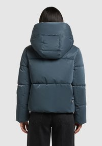 Paminkštinta mėlyna striukė su aukštu apykakliu ir gobtuvu. Medžiaga blizgi, su siūtu raštu. Mažas logotipo detalė apatinėje pusėje.