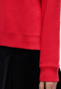 Κοντινή λήψη από το μανίκι και το κάτω μέρος ενός κόκκινου μακρυμάνικου πουκάμισου πάνω από μαύρο παντελόνι κορντόνι, εμφανίζοντας την υφή του υφάσματος και τις λεπτομέρειες ραφής.