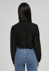 Chemise noire texturée à manches longues avec un col froncé, associée à un jean en denim bleu clair taille haute. La vue arrière montre des détails froncés.