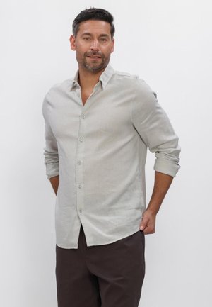 Mann mit kurzem dunklem Haar und Bart, trägt ein hellgraues Hemd mit hochgekrempelten Ärmeln und dunkle Hose, steht vor weißem Hintergrund.