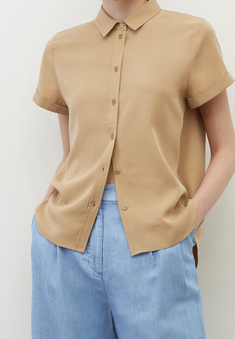 Personne portant une chemise beige à manches courtes boutonnée, partiellement rentrée dans un pantalon bleu clair, mains dans les poches, devant un fond uni.