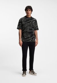 Νεαρός άνδρας που φοράει μαύρο και γκρι μπλουζάκι με αφηρημένο σχέδιο, μαύρο παντελόνι και λευκά αθλητικά παπούτσια με πράσινες και καφέ λεπτομέρειες, στέκεται σε απλό φόντο.