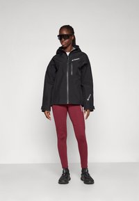 Schwarze wasserdichte Jacke mit Reißverschluss, ausgestattet mit einer abnehmbaren Kapuze und einem Logo. Getragen über burgunderfarbenen Leggings und schwarzen Sportschuhen.