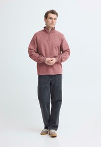 Trui in stofroze, met een halve ritssluiting bij de kraag, geribbelde boorden en een ronde accentdetail op de borst; gecombineerd met een donkere spijkerbroek.