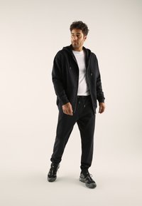 Black zip-up hoodie with a drawstring hood, paired with matching black sweatpants. The model wears a white t-shirt underneath and athletic shoes.