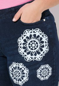 Vaqueros de mezclilla azul oscuro con tres estampados de encaje blanco en la zona de los muslos. El diseño presenta detalles geométricos intrincados.