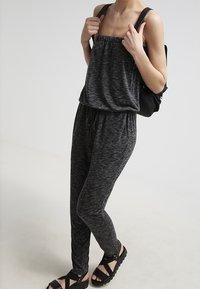 Jumpsuit nero melange con scollo elasticizzato e vita con cordoncino, abbinato a sandali neri. Design semplice, vestibilità rilassata, materiale comodo.