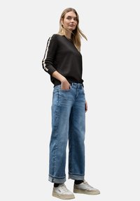 Czarny sweter z dzianiny z kremowymi paskami na rękawach, w połączeniu z jasnoniebieskimi spodniami o szerokim kroju i beżowymi trampkami. Gładka faktura, casualowy fason.